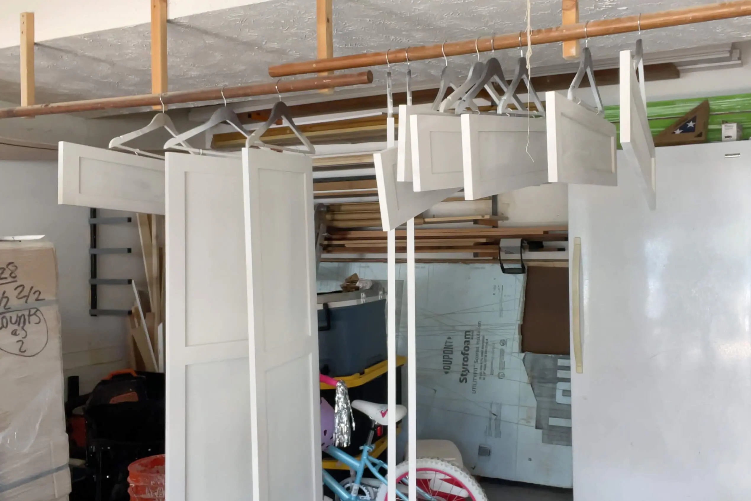 Multiple painted cabinet doors hanging from wooden rods on hangers in a garage workshop, with tools, supplies, and a child’s bicycle visible in the background.
